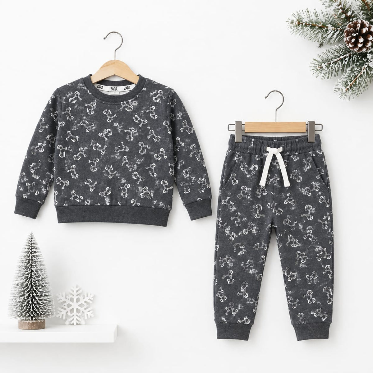 A children's two-piece outfit set consisting of a long-sleeve crewneck sweatshirt and matching jogger pants, both in dark gray with a white snowman pattern, hanging on wooden hangers against a white wall with holiday decorations.