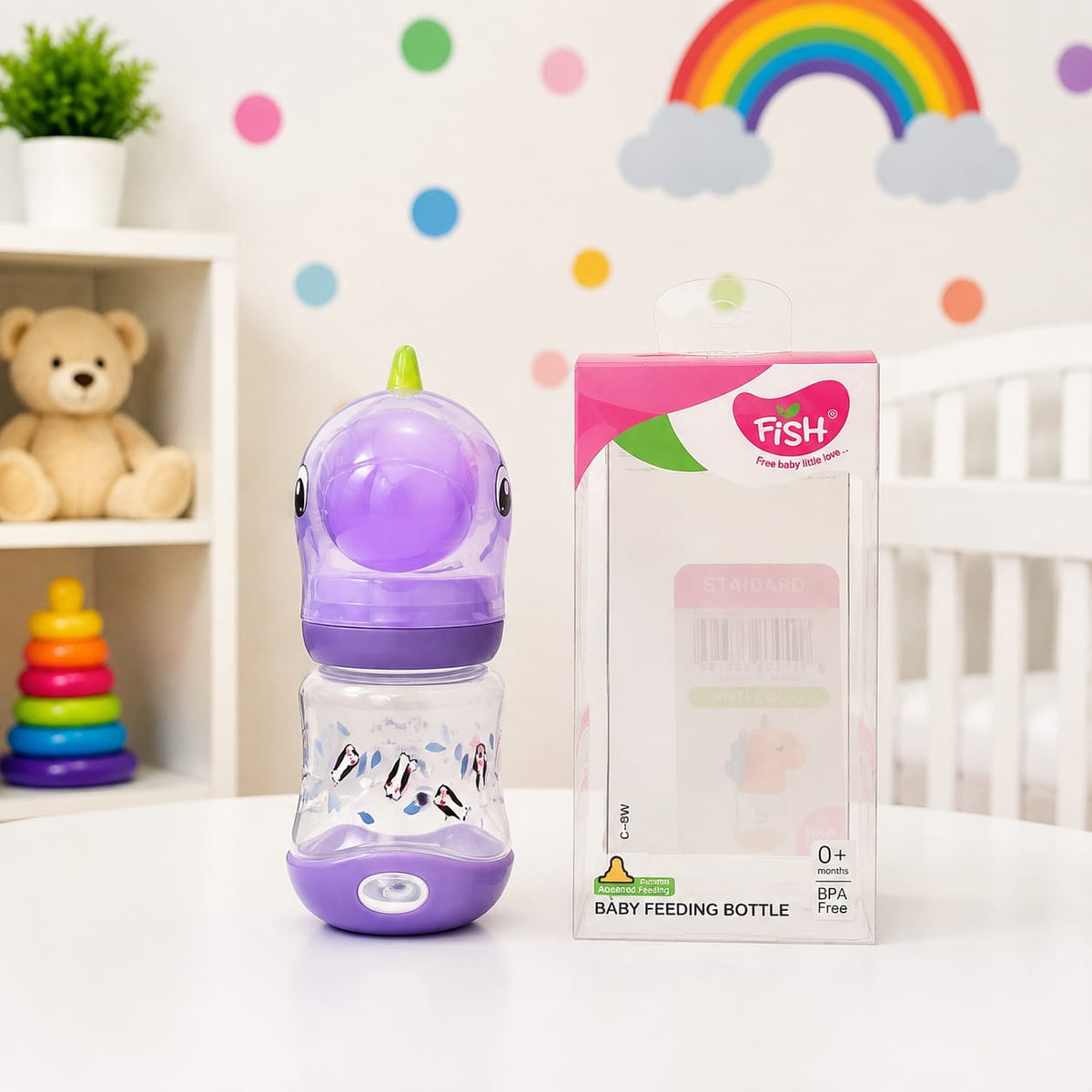 A purple and clear baby feeding bottle with a dinosaur design stands next to its retail box on a white table. The background shows a soft, decorated nursery setting with a teddy bear, rainbow, clouds, and colorful dots.