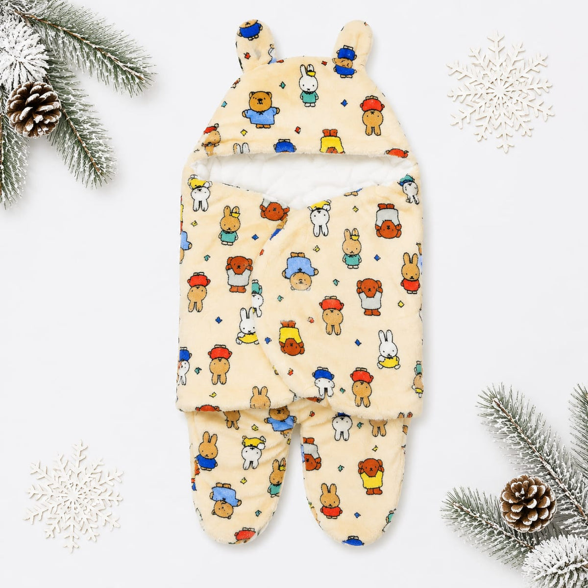 A baby footed swaddle blanket with a hood, featuring a playful multicolor cartoon animal print on a warm cream background. The swaddle is styled in a flat-lay arrangement with winter-themed props including pine branches, pinecones, and white snowflakes on