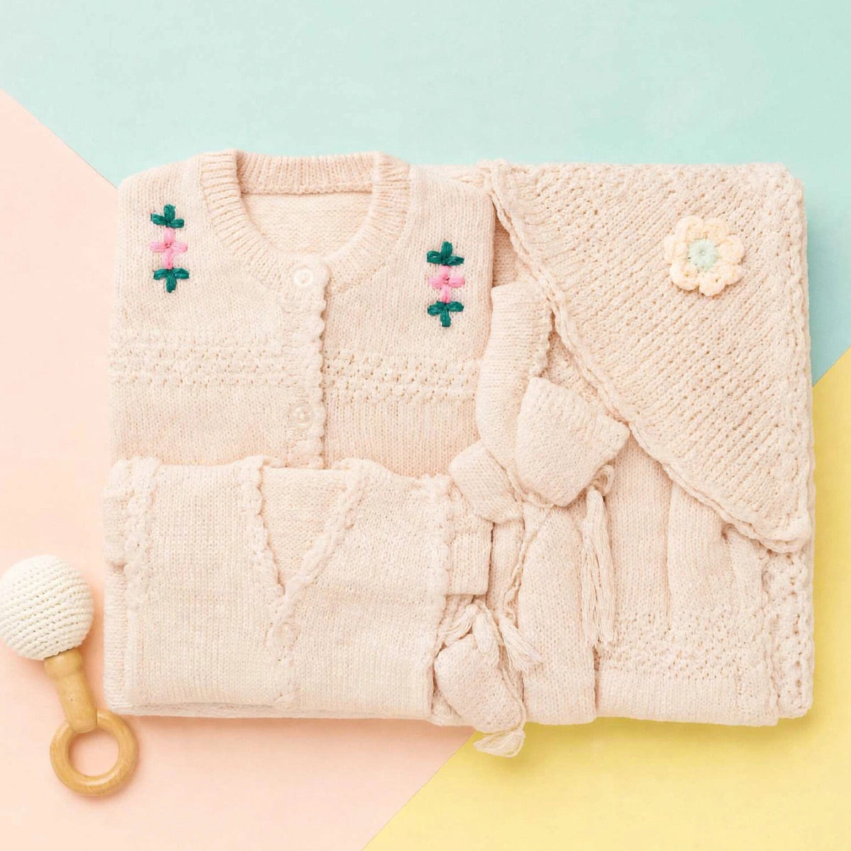 A folded knit baby outfit set in a soft blush pink, including a cardigan with embroidered flowers, a matching wrap or blanket with a flower applique, and a small wooden-handled baby rattle on a pastel geometric background.