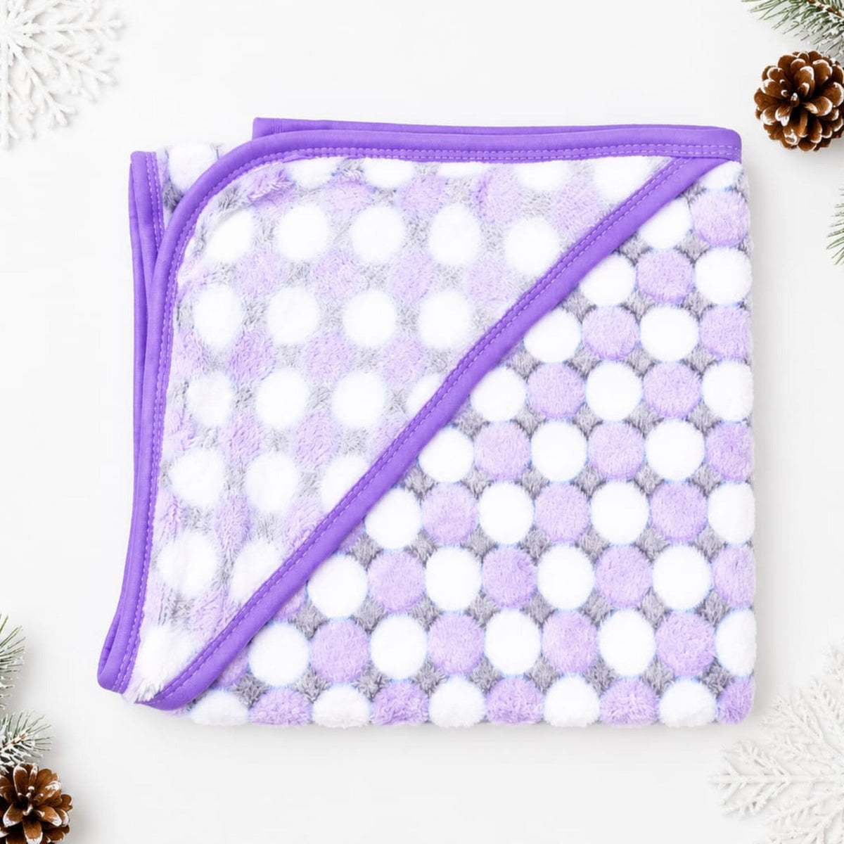 A folded plush blanket with a purple and white polka dot pattern and solid purple trim, displayed on a white background with decorative pinecones and snowflakes.