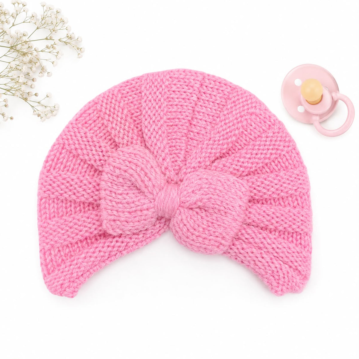 A pink knitted baby turban hat with a large matching bow, displayed on a white background with a pacifier and small white flowers as props.