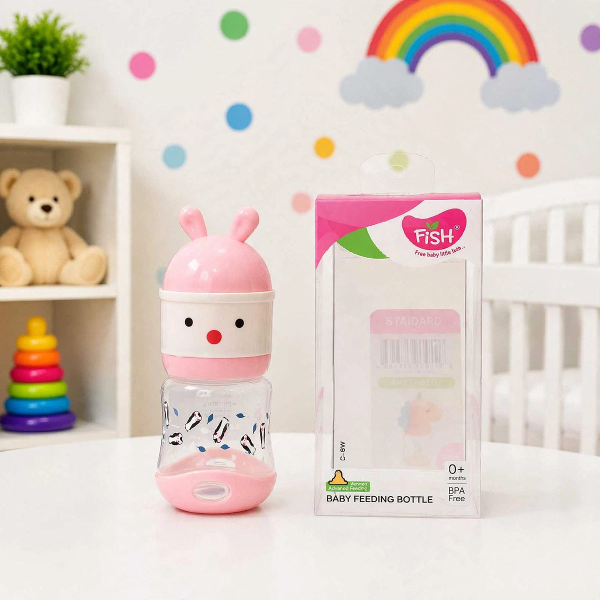 A pink and white baby feeding bottle with a bunny design next to its retail packaging box, set on a white table in a decorated nursery room.