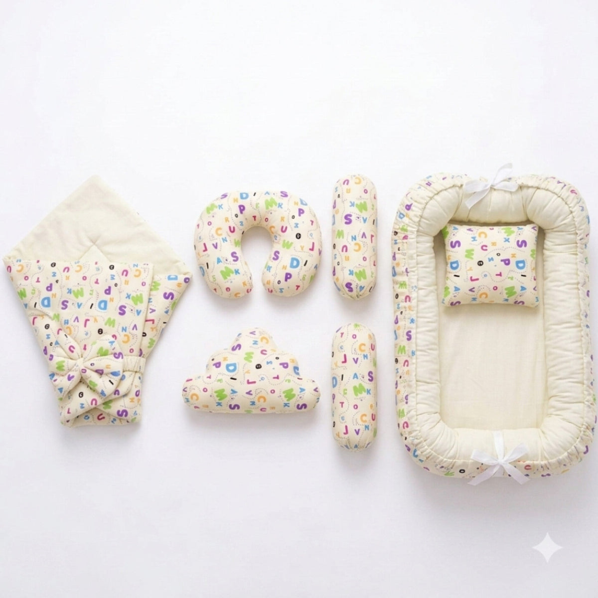 A baby bedding set laid out on a white background, including a quilted blanket with a colorful alphabet print and bow, a U-shaped pillow, two cylindrical pillows, a cloud-shaped pillow, and a bassinet cover with matching printed fabric and white ribbon ti