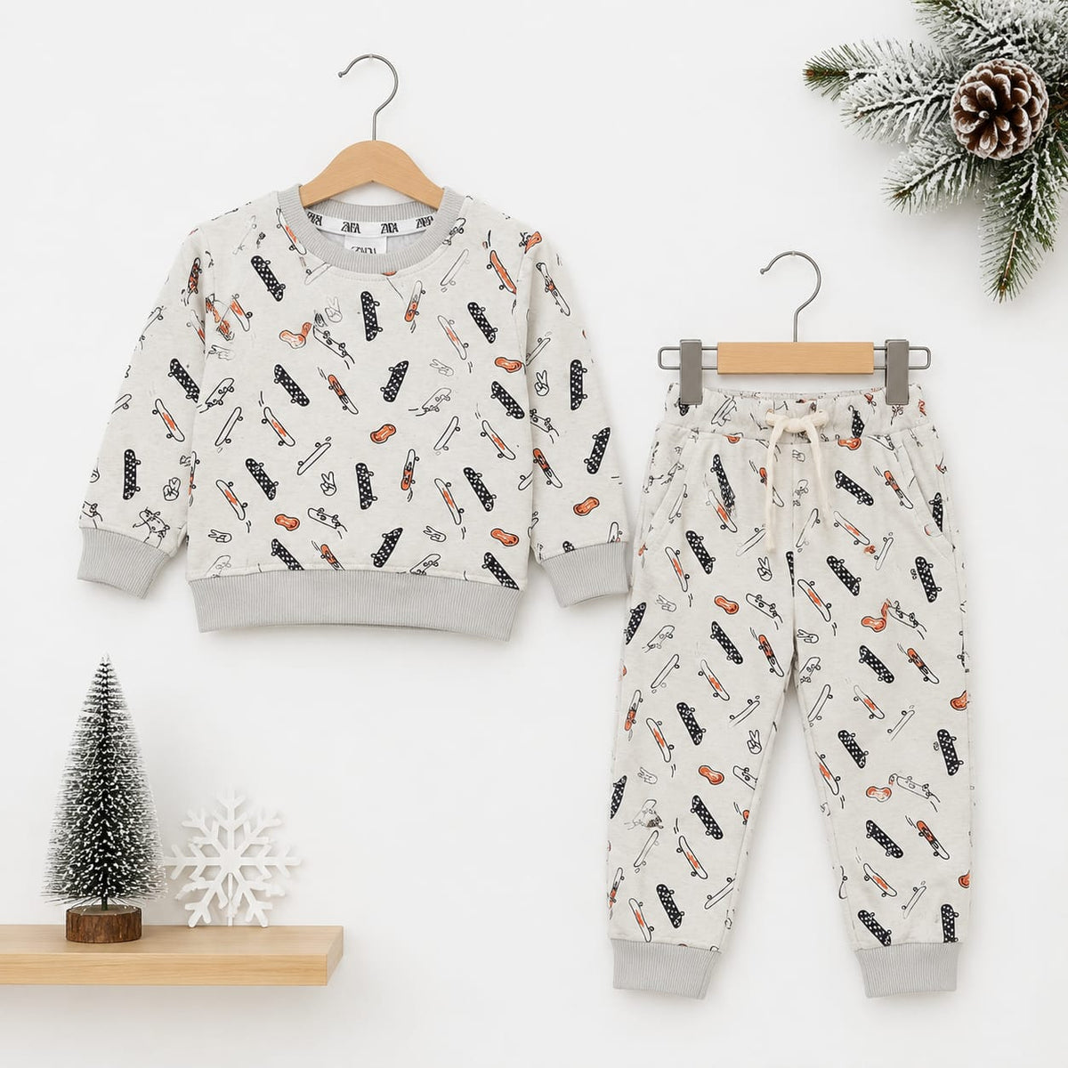A children's two-piece clothing set featuring a long-sleeve sweatshirt and matching jogger pants, both in a warm off-white color with a playful print of black and orange winter-themed illustrations. The set is displayed on wooden hangers against a clean w