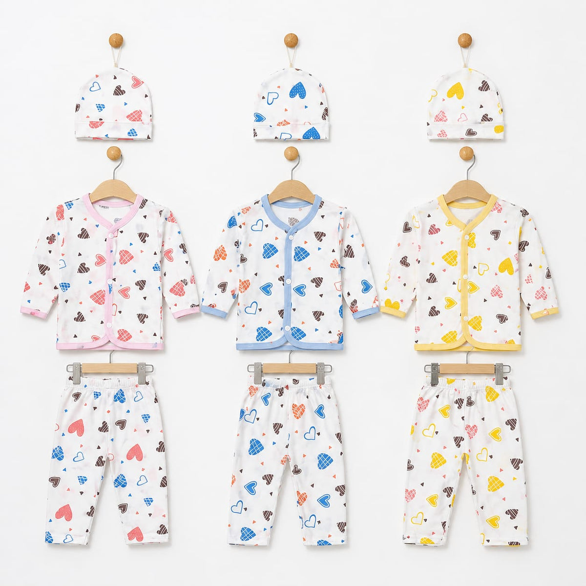Three baby clothing sets hanging on wooden hangers against a white wall, each set including a hat, cardigan, and pants. The sets are white with colorful heart and cloud prints in pink, blue, brown, and yellow, with different trim colors (pink, blue, and y