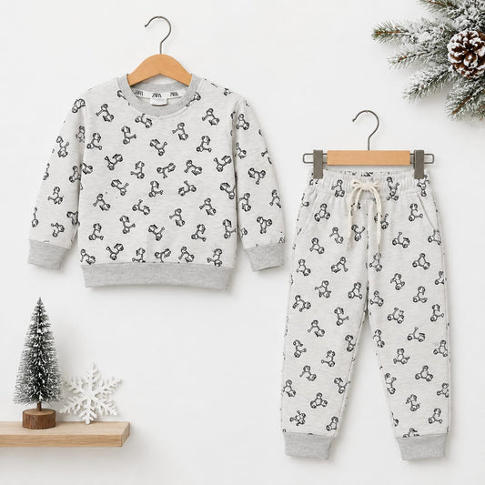 A children's two-piece pajama set hanging on wooden hangers against a white wall. The set includes a long-sleeve sweatshirt and matching jogger pants in a light gray fabric with a black repeating graphic pattern, styled with small holiday props including 