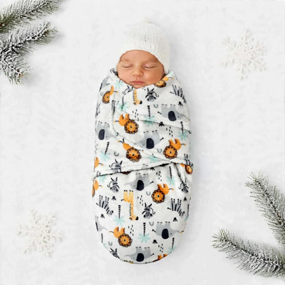 A sleeping newborn baby swaddled in a white blanket with a colorful safari animal print, wrapped in a soft white knit hat, laid flat on a white textured surface with snowy decorative props.