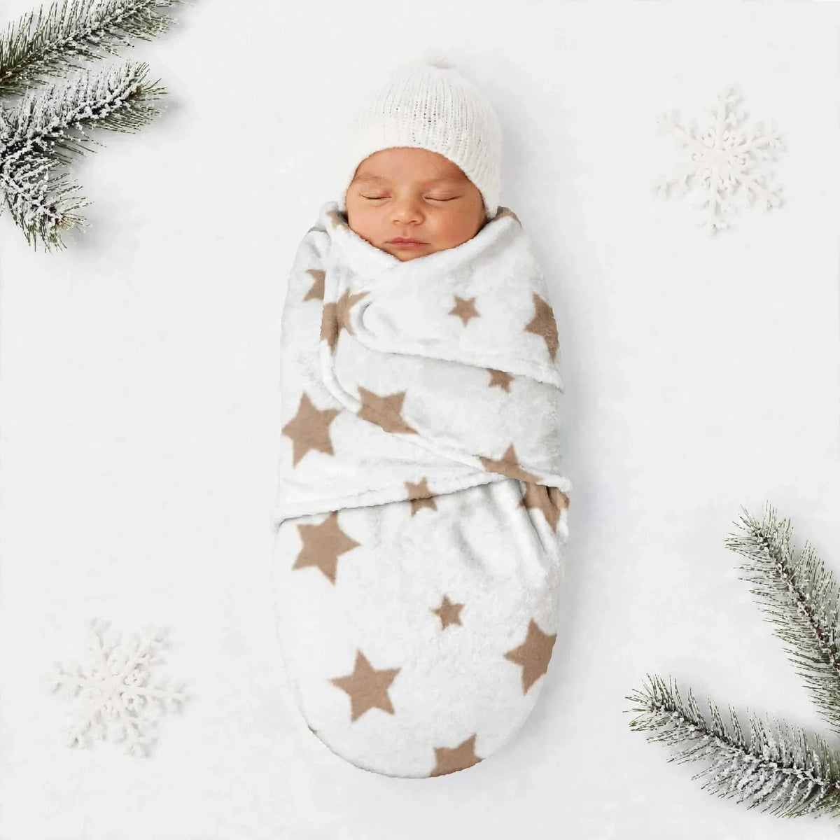A sleeping newborn baby swaddled in a white fleece blanket with brown star patterns, wearing a matching white knit beanie, laid flat on a white background with decorative snowflakes and pine branches.