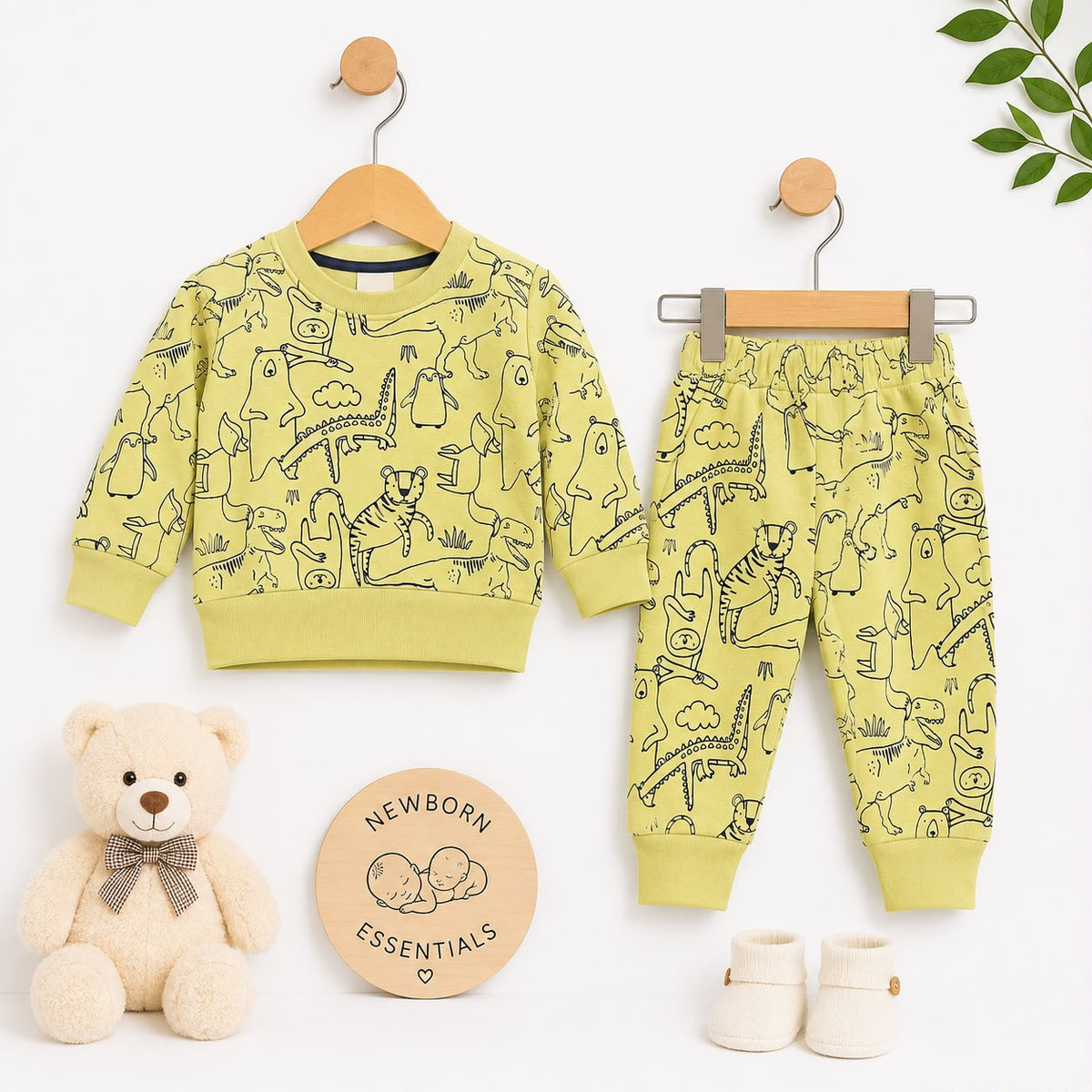 A baby clothing set displayed on wooden hangers against a white background, including a long-sleeve sweatshirt and matching jogger pants in a pale yellow-green color with black animal line drawings. A teddy bear, a round 'Newborn Essentials' card, and cre