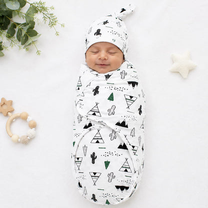 A sleeping newborn baby swaddled in a white muslin swaddle blanket with a black and green geometric teepee and cactus pattern, wearing a matching beanie. The baby is photographed on a white fabric background with baby props including a wooden star teether