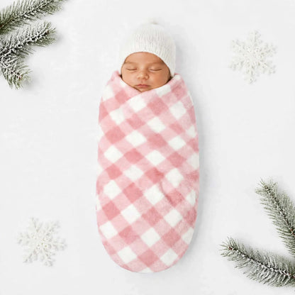 A sleeping newborn baby swaddled in a pink and white gingham blanket wrap, wearing a white knit beanie, laid flat on a white background with decorative snowy snowflakes and pine branches in the corners.