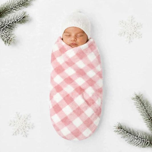 A sleeping newborn baby swaddled in a pink and white gingham blanket wrap, wearing a white knit beanie, laid flat on a white background with decorative snowy snowflakes and pine branches in the corners.