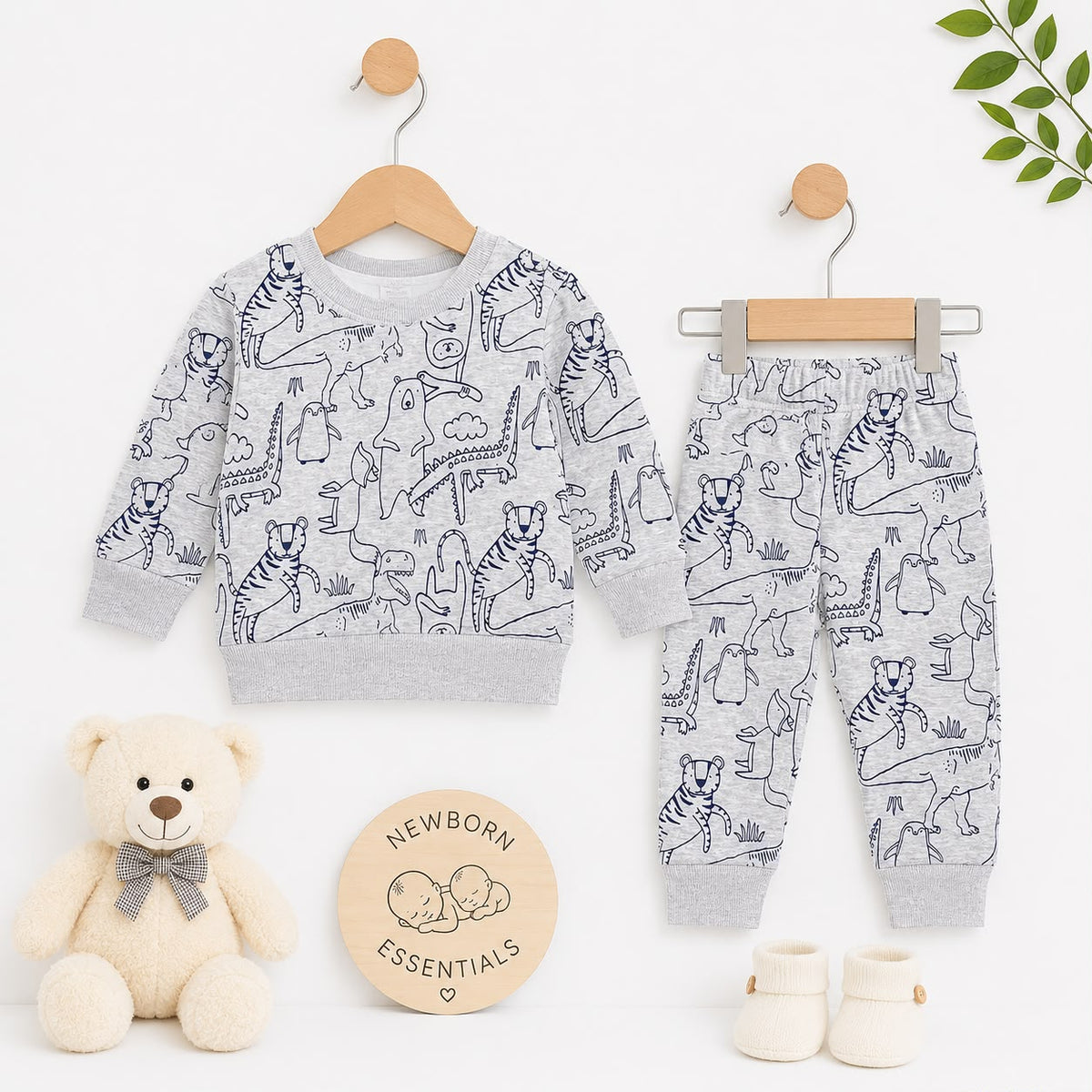 A baby clothing set displayed on wooden hangers against a clean white background, styled with a cream teddy bear, a round 'Newborn Essentials' sign, and small cream booties. The set includes a long-sleeve sweatshirt and matching pants in light heather gra