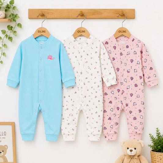 Three baby rompers in blue, white, and pink hanging on a wooden rack with a teddy bear and plants in the background.