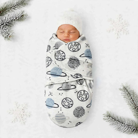 A sleeping newborn baby swaddled in a white wrap blanket with a blue and black space-themed print of planets, moons, and stars, wearing a white knit hat. The baby is photographed from a top-down flat-lay perspective on a white textured background decorate