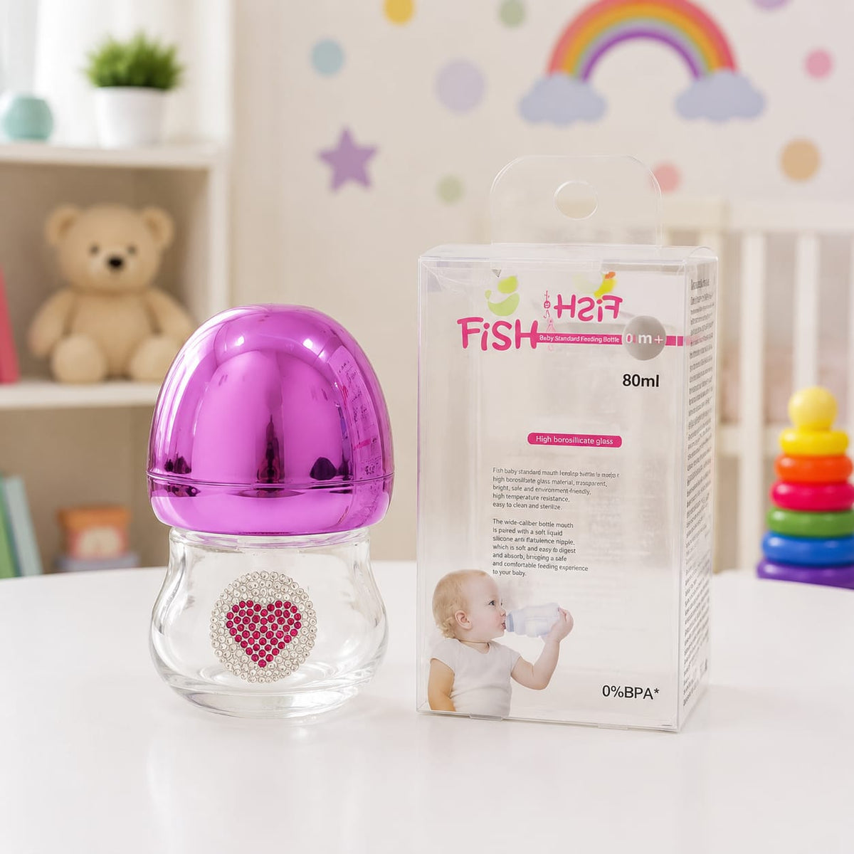 A clear baby bottle with a shiny metallic magenta-purple cap and a rhinestone heart emblem on the front, shown next to its retail box labeled 'Her Fish' on a white tabletop. The background is a softly blurred nursery setting with a teddy bear, shelf, and 