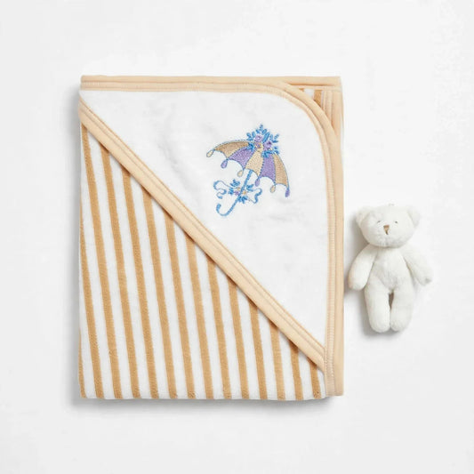 A folded baby hooded blanket with alternating cream and tan stripes, featuring a white hooded section with a purple and blue embroidered umbrella design. A small white plush teddy bear sits to the right on a clean white background.