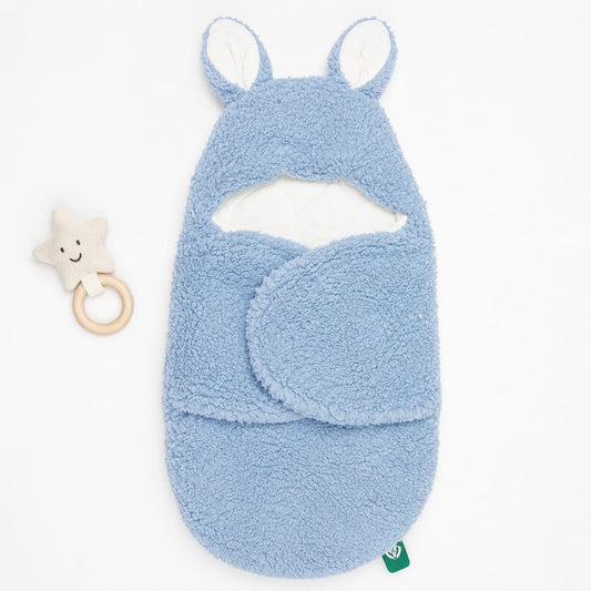 A light blue plush baby swaddle blanket with bunny ears and a matching footed cowl, shown on a white background next to a small star-shaped wooden teether.