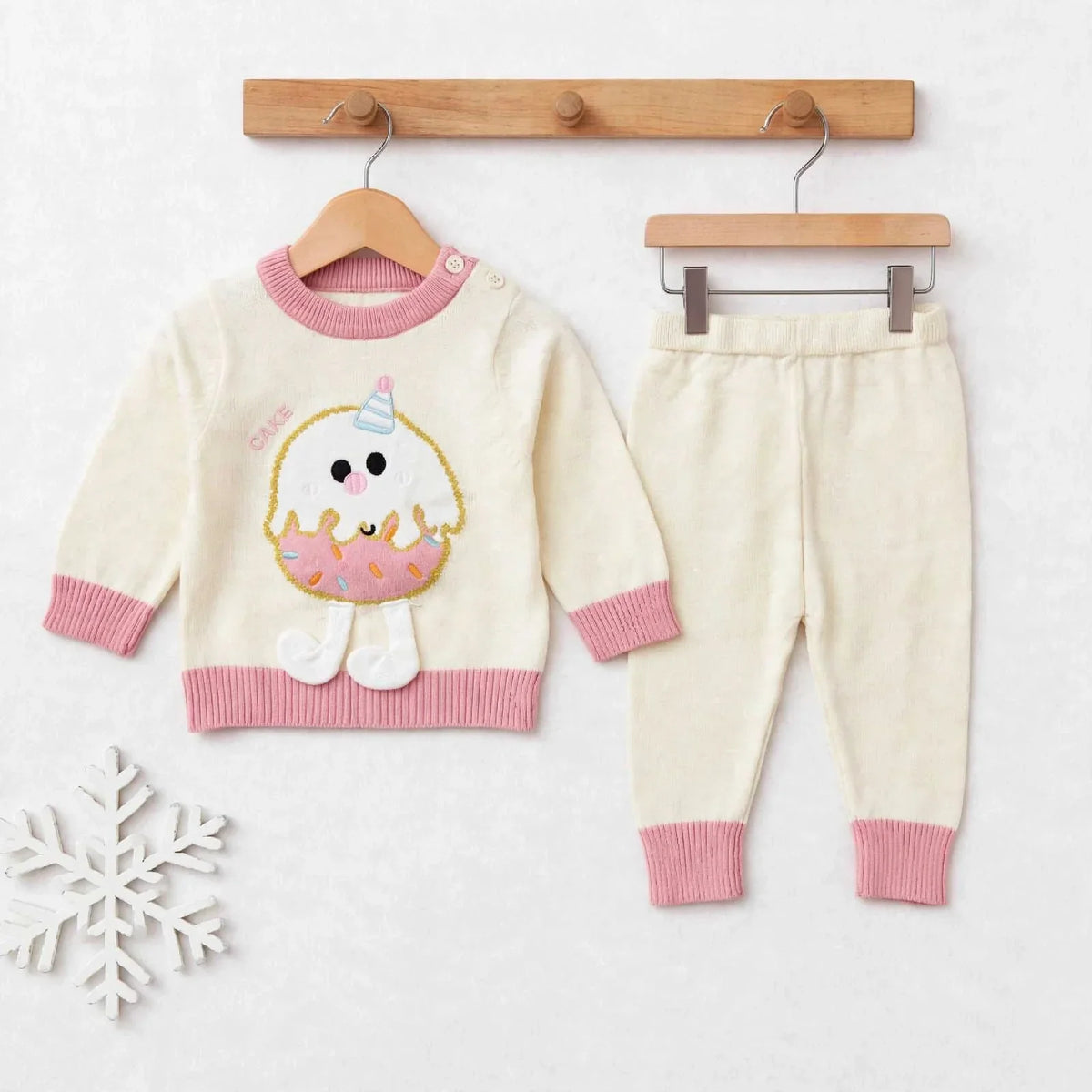 A baby knit two-piece outfit set consisting of a cream sweater with pink ribbed trim and a cute donut character applique, paired with matching cream pants with pink ribbed cuffs, displayed on wooden hangers against a light background.