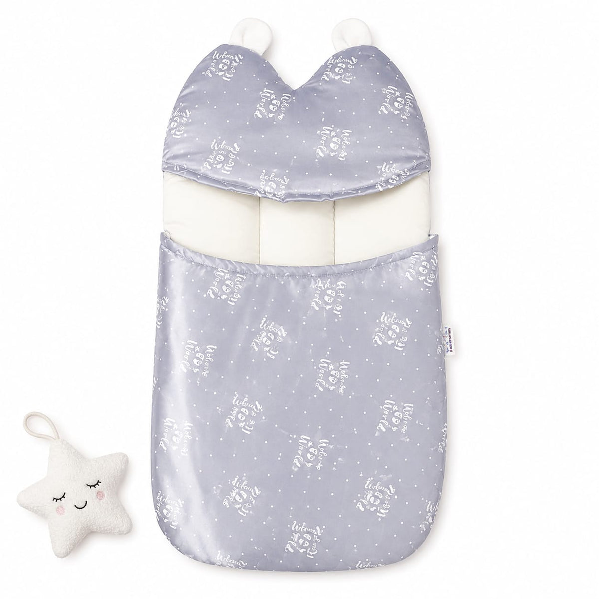 A baby footmuff in a light lavender-gray satin-like fabric with a white floral and star pattern, featuring a white inner lining and two white pom-poms. A small white plush star toy with embroidered facial features is placed to the left on a clean white ba