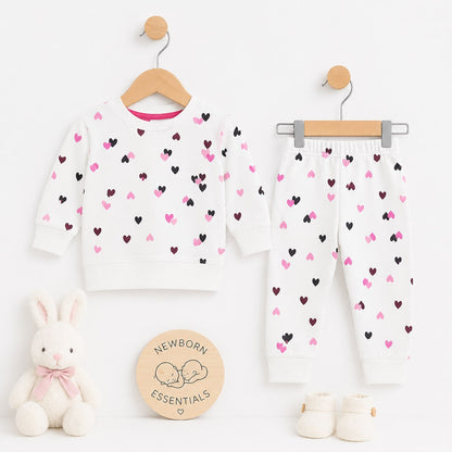 A baby clothing set displayed on wooden hangers against a white background, featuring a long-sleeve top and matching pants with an allover heart pattern in pink and black. The set is styled with a plush white bunny toy, a round wooden 'Newborn Essentials'