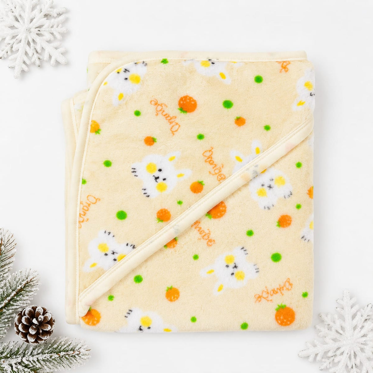 A folded cream-colored hooded baby blanket with a daisy and orange pattern, decorated with 'Daisy' text and small green dots, styled with pine branches, a pinecone, and white snowflakes on a white background.