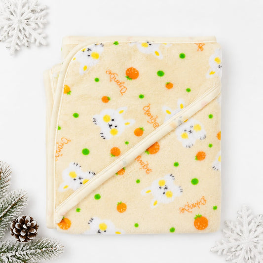 A folded cream-colored hooded baby blanket with a daisy and orange pattern, decorated with 'Daisy' text and small green dots, styled with pine branches, a pinecone, and white snowflakes on a white background.