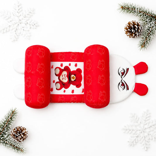 A red and white baby head and neck support pillow styled in a flat lay with white snowflakes, pinecones, and green pine branches on a white background.