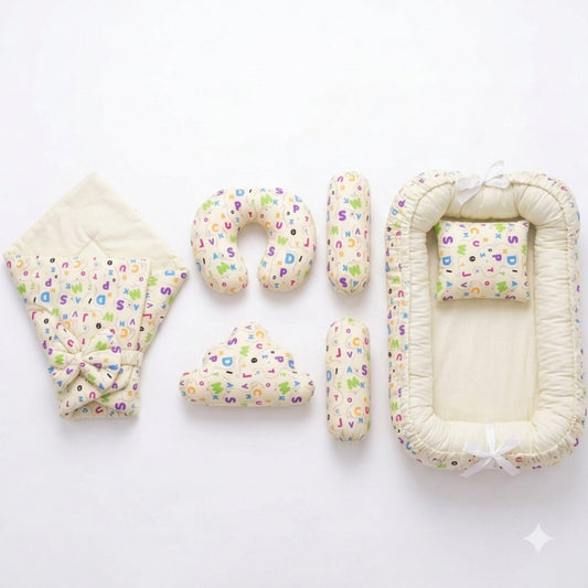 A baby bedding set laid out on a white background, including a quilted blanket with a colorful alphabet print and bow, a U-shaped pillow, two cylindrical pillows, a cloud-shaped pillow, and a bassinet cover with matching printed fabric and white ribbon ti