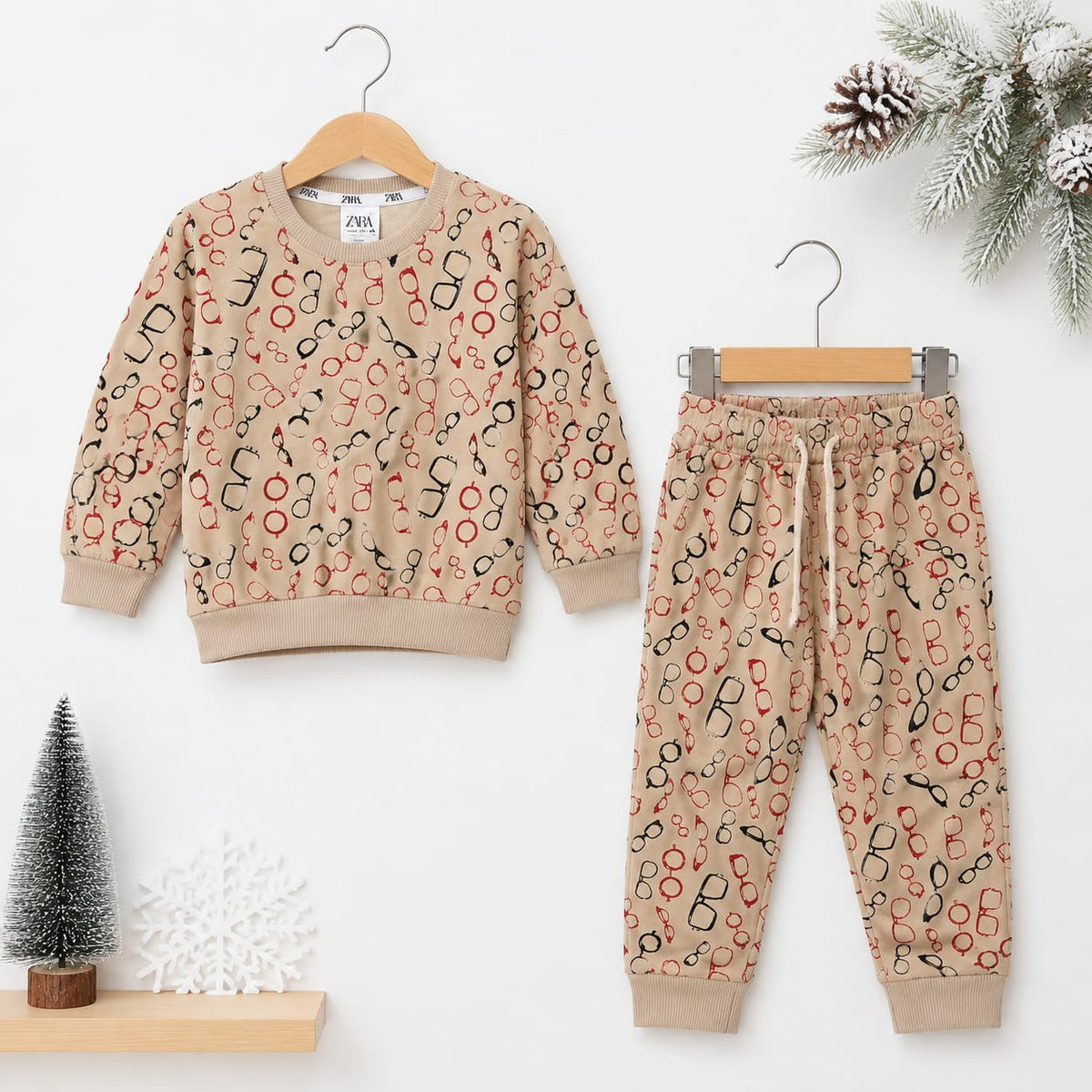 A children's two-piece pajama set hanging on wooden hangers against a white background, featuring a beige sweatshirt and matching joggers with an all-over abstract print in black, red, and orange. Decorative holiday props including a small tree, snowflake