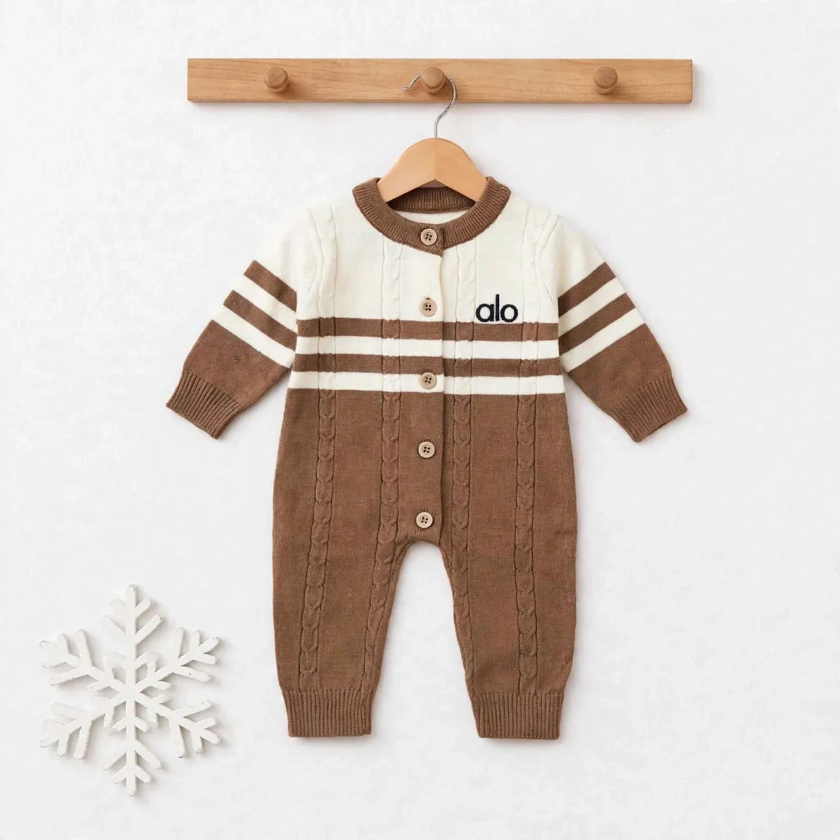 A baby knit cardigan jumpsuit in brown and cream, hanging on a wooden hanger from a wooden peg rack. The jumpsuit features a cream upper chest and shoulders with brown stripes, a brown button-front closure, and a brown cable-knit lower body.