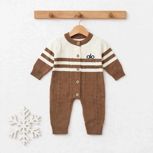 A baby knit cardigan jumpsuit in brown and cream, hanging on a wooden hanger from a wooden peg rack. The jumpsuit features a cream upper chest and shoulders with brown stripes, a brown button-front closure, and a brown cable-knit lower body.