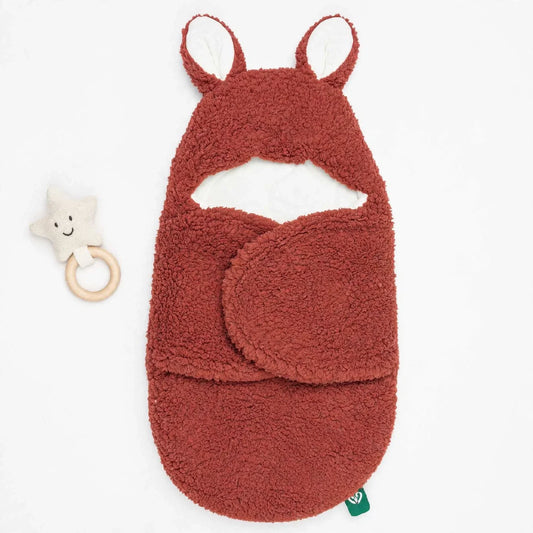 A terracotta-colored plush baby wrap or swaddle with a hood featuring pointed ears, shown on a white background next to a small cream star rattle.