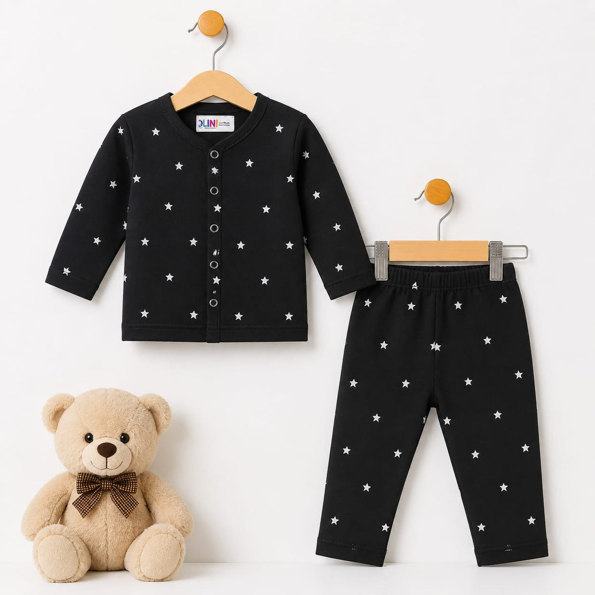 A baby clothing set consisting of a black long-sleeve cardigan top and matching pants, both decorated with small white star patterns. The set is displayed on wooden hangers next to a beige teddy bear toy on a plain white background.